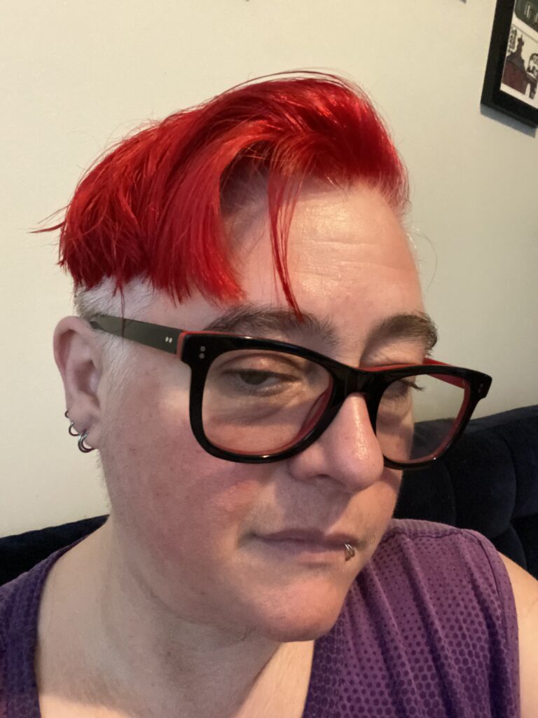 Selfie of K Lucian. They have bright dyed red short hair and are wearing glasses with black and red frames